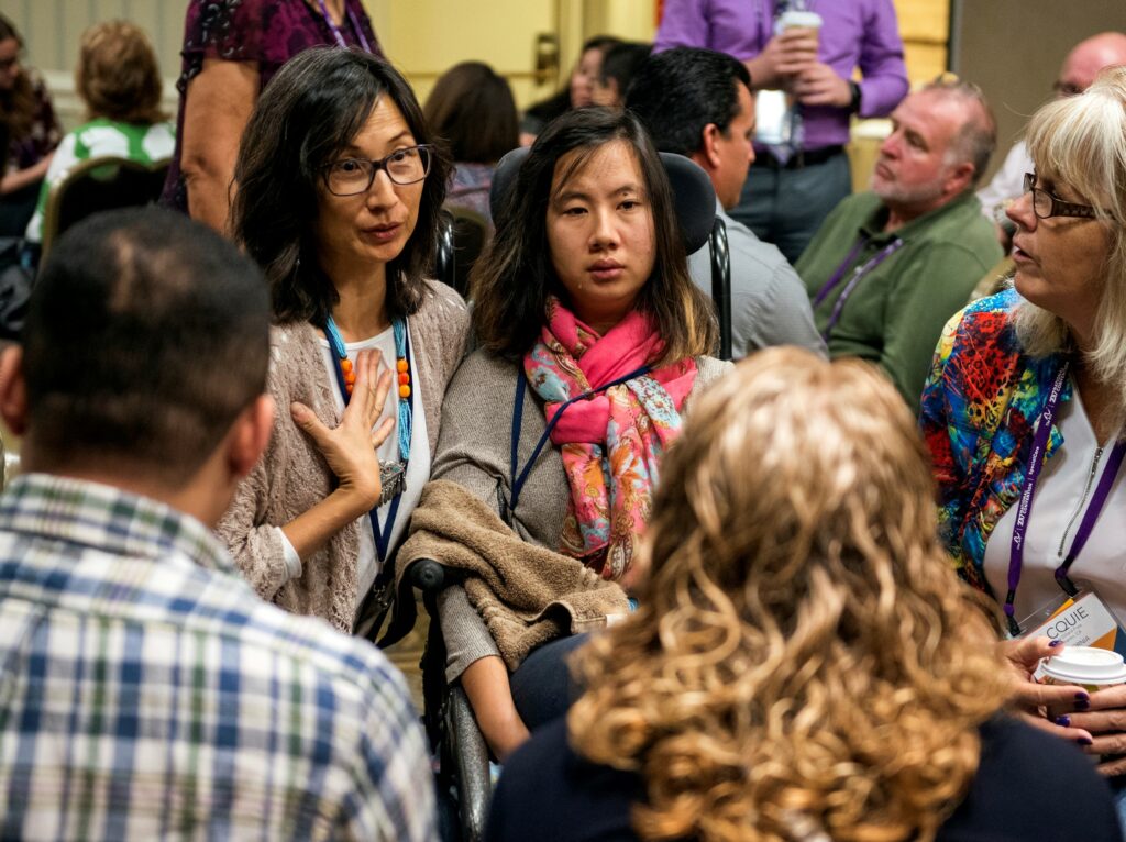 A woman caught mid-sentence in a serious chat with three adults in a crowded room crouches beside a young woman in a wheelchair.